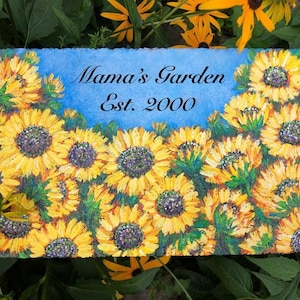 May include: A painted stone sign with a blue background and a cluster of yellow sunflowers. The sign reads "Mama's Garden Est. 2000".