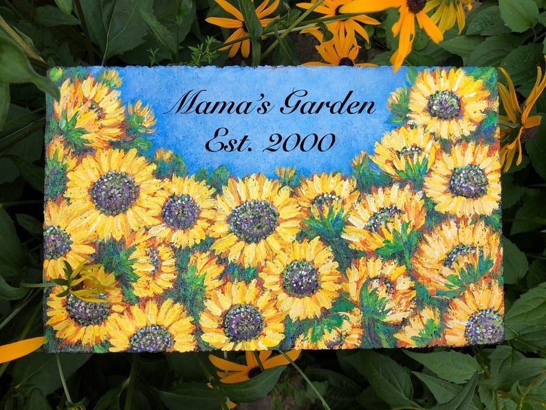 May include: A painted stone sign with a blue background and a cluster of yellow sunflowers. The sign reads "Mama's Garden Est. 2000".