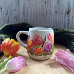 May include: A white ceramic mug with a floral design featuring pink, purple, and orange tulips. The mug has a curved handle and is surrounded by fresh tulips in various colors. The background includes a green knitted scarf and a wooden surface.