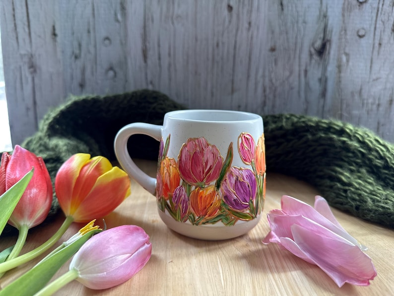 May include: A white ceramic mug with a floral design featuring pink, purple, and orange tulips. The mug has a curved handle and is surrounded by fresh tulips in various colors. The background includes a green knitted scarf and a wooden surface.