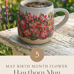 May include: A ceramic mug featuring hand-painted pink hawthorn flowers with green leaves on a cream-colored base. The mug contains a dark liquid and includes text that reads: 5 MAY BIRTH MONTH FLOWER Hawthorn Mug HAND PAINTED WITH LOVE.