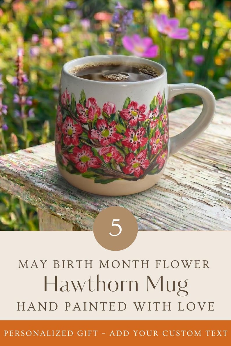 May include: A ceramic mug featuring hand-painted pink hawthorn flowers with green leaves on a cream-colored base. The mug contains a dark liquid and includes text that reads: 5 MAY BIRTH MONTH FLOWER Hawthorn Mug HAND PAINTED WITH LOVE.