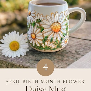 May include: A ceramic mug featuring a hand-painted daisy floral design with white petals and yellow centers, accompanied by a single fresh daisy. The text on the image reads: 4 APRIL BIRTH MONTH FLOWER Daisy Mug HAND PAINTED WITH LOVE.