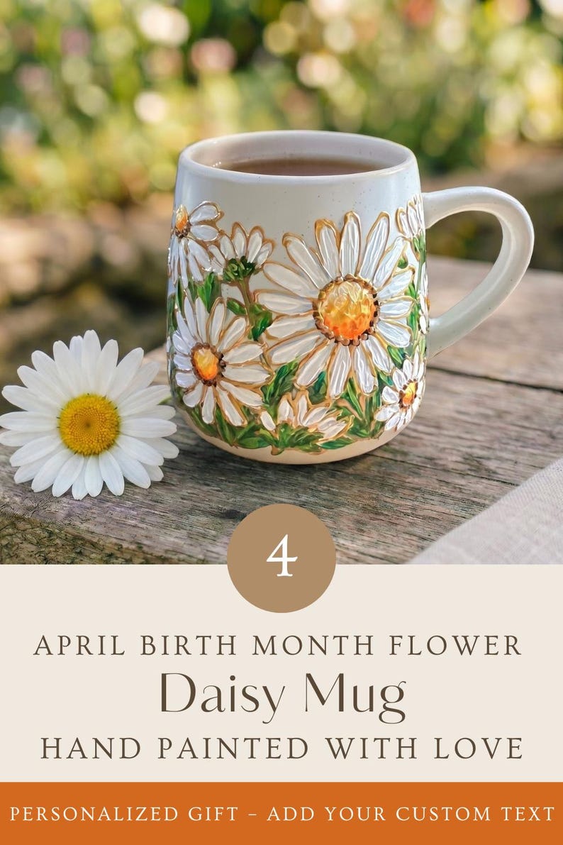 May include: A ceramic mug featuring a hand-painted daisy floral design with white petals and yellow centers, accompanied by a single fresh daisy. The text on the image reads: 4 APRIL BIRTH MONTH FLOWER Daisy Mug HAND PAINTED WITH LOVE.