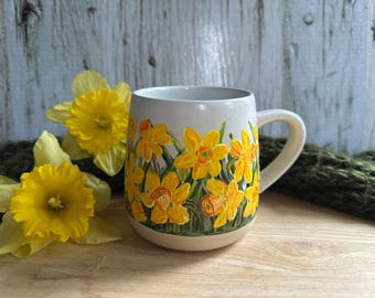 Könnte beinhalten: Eine Keramiktasse mit cremefarbenem Boden und Henkel, verziert mit einem handgemalten Design aus gelben Narzissen. Die Blumen sind vor einem hellen Hintergrund angeordnet. Die Tasse wird mit echten Narzissen präsentiert.