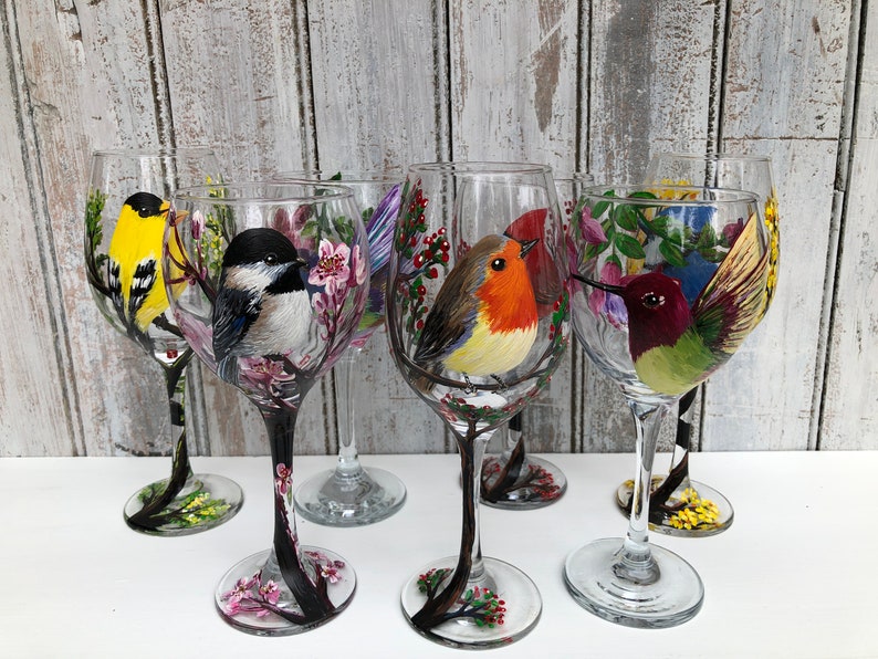 May include: Six clear wine glasses, each hand-painted with a different colorful bird design. The birds include a yellow finch, a black-capped chickadee, a robin, and a hummingbird. The stems are painted black with floral accents.