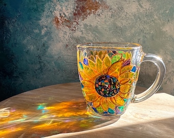 May include: A glass mug featuring a hand-painted sunflower design. The sunflower is yellow with a brown centre and green leaves. The mug has a clear glass handle.