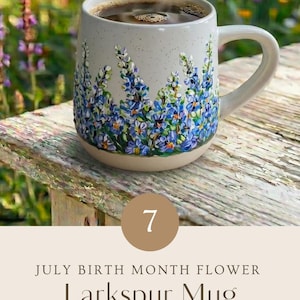 May include: A speckled ceramic mug featuring a hand-painted design of blue and purple larkspur flowers around the base. The mug contains a dark liquid and includes text that reads 7 JULY BIRTH MONTH FLOWER Larkspur Mug HAND PAINTED WITH LOVE.