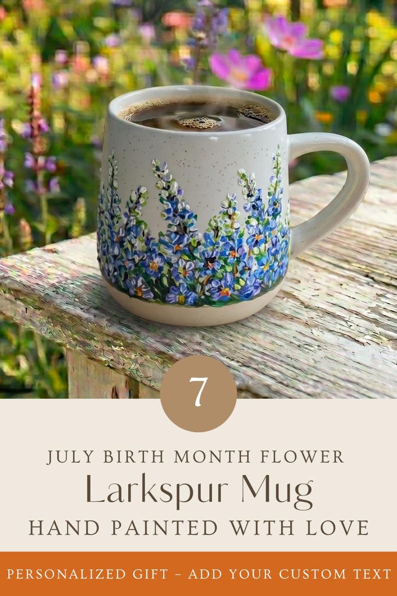 May include: A speckled ceramic mug featuring a hand-painted design of blue and purple larkspur flowers around the base. The mug contains a dark liquid and includes text that reads 7 JULY BIRTH MONTH FLOWER Larkspur Mug HAND PAINTED WITH LOVE.
