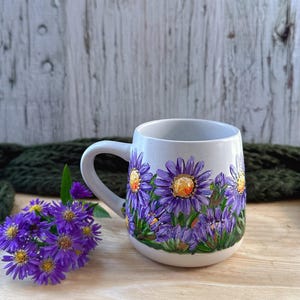 May include: A ceramic mug with a white base and a curved handle. The mug is decorated with hand-painted purple flowers with yellow centers and green leaves. The mug is sitting on a wooden surface.