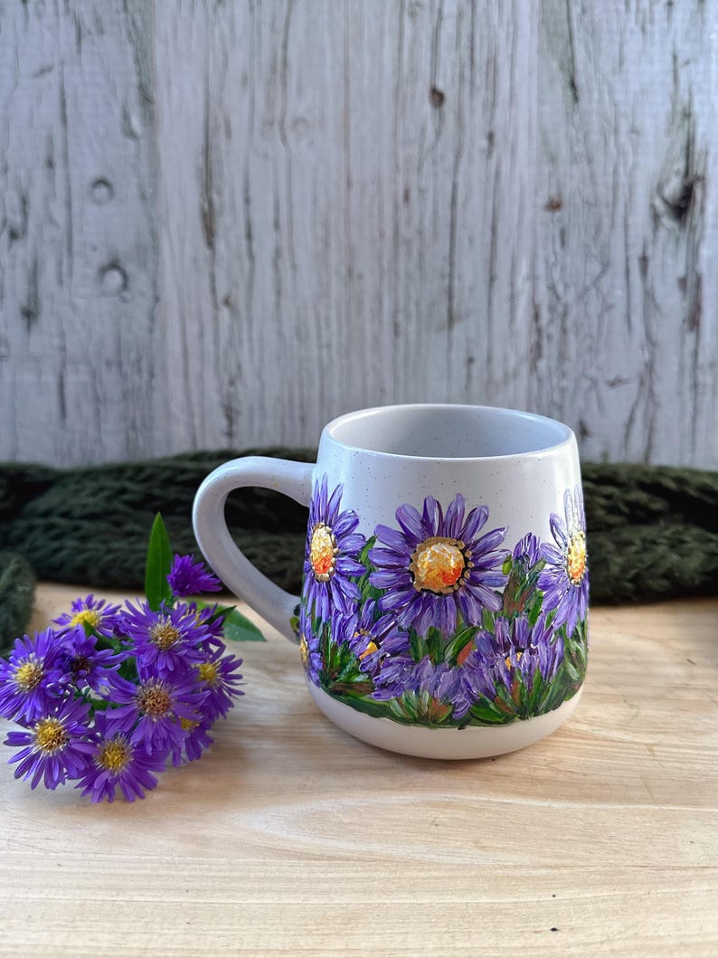 May include: A ceramic mug with a white base and a curved handle. The mug is decorated with hand-painted purple flowers with yellow centers and green leaves. The mug is sitting on a wooden surface.