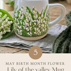 May include: A ceramic mug featuring a hand-painted design of white lily of the valley flowers with green leaves on a light-colored background. The text on the image reads: 5 MAY BIRTH MONTH FLOWER Lily of the valley Mug HAND PAINTED WITH LOVE.