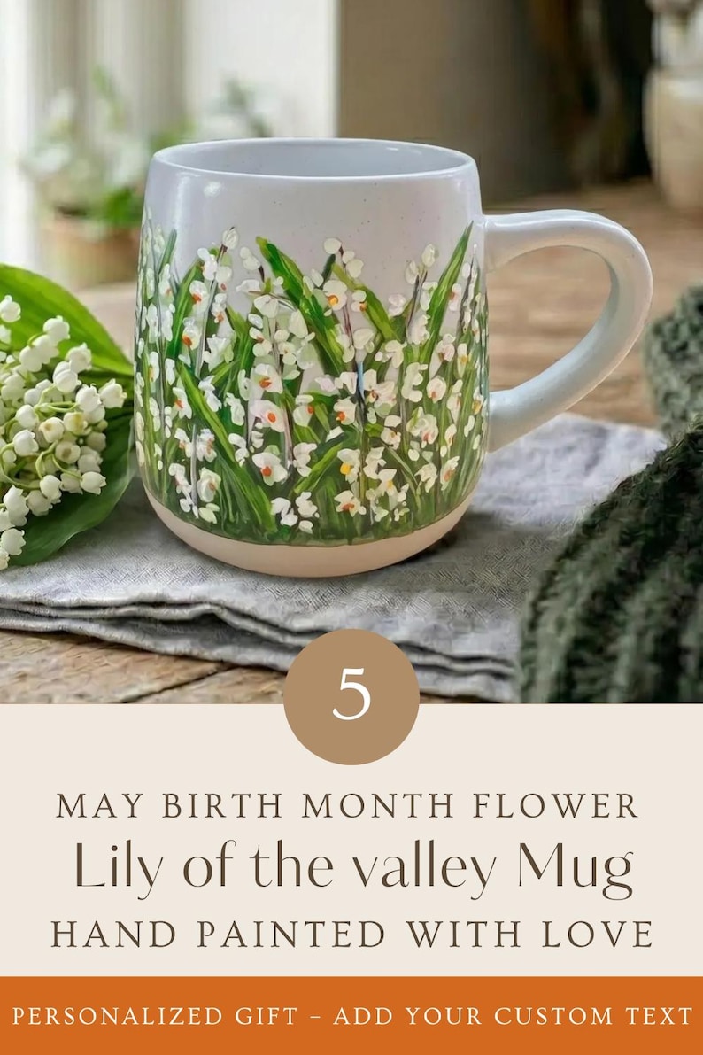 May include: A ceramic mug featuring a hand-painted design of white lily of the valley flowers with green leaves on a light-colored background. The text on the image reads: 5 MAY BIRTH MONTH FLOWER Lily of the valley Mug HAND PAINTED WITH LOVE.