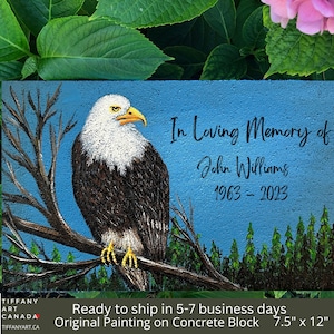 May include: A hand-painted concrete block memorial with a bald eagle perched on a branch. The background is a blue sky with a forest of green trees. The text reads "In Loving Memory of John Williams 1963-2023".