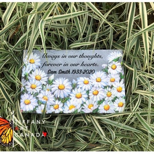 May include: A gray stone memorial with a painted design of white daisies. The stone has the text "Always in our thoughts, forever in our hearts. Sam Smith 1933-2020".