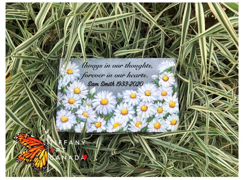 May include: A gray stone memorial with a painted design of white daisies. The stone has the text "Always in our thoughts, forever in our hearts. Sam Smith 1933-2020".