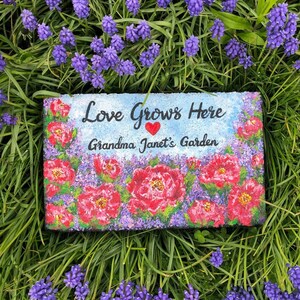 May include: A painted stone with the text "Love Grows Here Grandma Janet's Garden" and a red heart. The stone is surrounded by purple flowers.