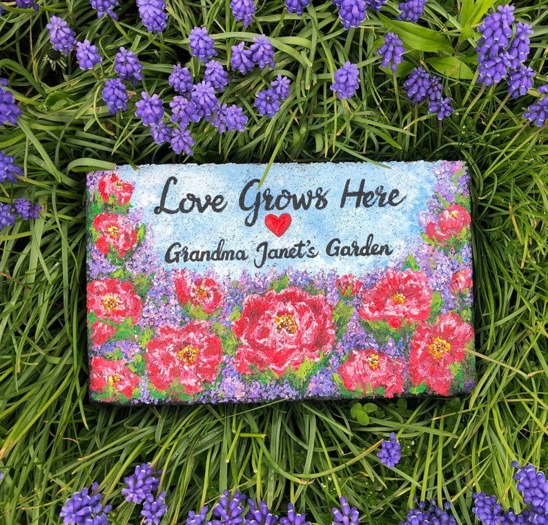 May include: A painted stone with the text "Love Grows Here Grandma Janet's Garden" and a red heart. The stone is surrounded by purple flowers.