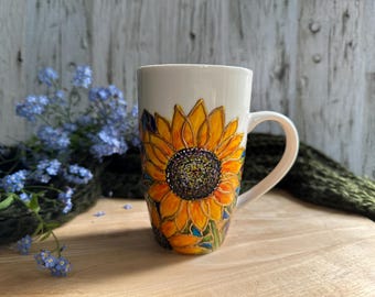 May include: White ceramic mug with a hand-painted sunflower design. The sunflower features yellow petals, a dark centre, and green and blue accents. The mug has a curved handle and is approximately 15 cm tall.