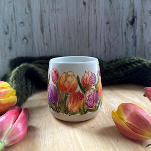 May include: A white ceramic mug with a hand-painted floral design featuring orange, purple, and pink tulips. The mug is surrounded by fresh tulips in various stages of bloom, with a green knitted scarf in the background.