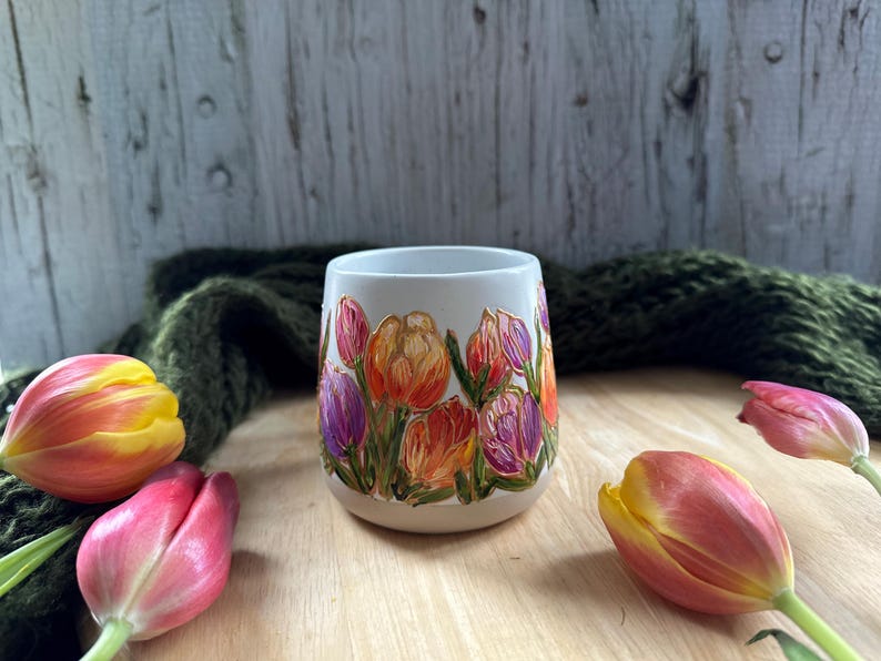 May include: A white ceramic mug with a hand-painted floral design featuring orange, purple, and pink tulips. The mug is surrounded by fresh tulips in various stages of bloom, with a green knitted scarf in the background.