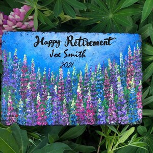 May include: A painted stone with a blue background and a field of purple and pink lupine flowers. The text "Happy Retirement Joe Smith 2021" is written on the stone.