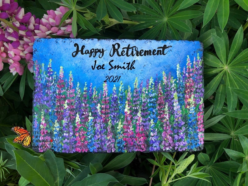 May include: A painted stone with a blue background and a field of purple and pink lupine flowers. The text "Happy Retirement Joe Smith 2021" is written on the stone.
