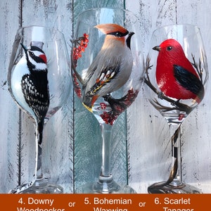 May include: Hand-painted wine glasses featuring bird designs: a Downy Woodpecker, a Bohemian Waxwing, and a Scarlet Tanager. Each glass showcases a detailed bird illustration with vibrant colors. The glasses are clear with a long stem.