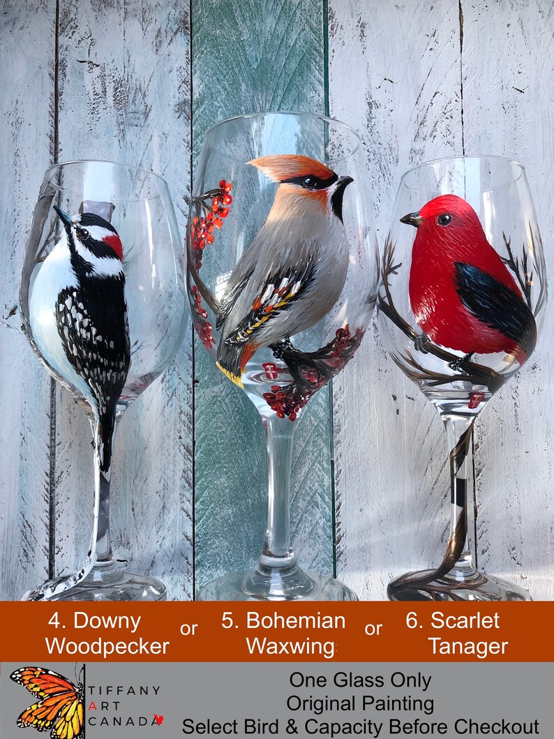 May include: Hand-painted wine glasses featuring bird designs: a Downy Woodpecker, a Bohemian Waxwing, and a Scarlet Tanager. Each glass showcases a detailed bird illustration with vibrant colors. The glasses are clear with a long stem.