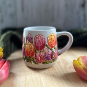 May include: A white ceramic mug adorned with a vibrant floral design featuring tulips in shades of pink, orange, and purple. The flowers are painted with textured brushstrokes, and the mug has a curved handle.