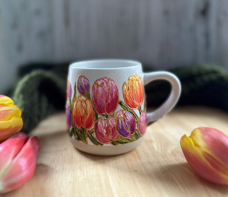 May include: A white ceramic mug adorned with a vibrant floral design featuring tulips in shades of pink, orange, and purple. The flowers are painted with textured brushstrokes, and the mug has a curved handle.