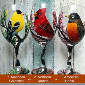 May include: Three hand-painted wine glasses, each featuring a different bird: an American Goldfinch, a Northern Cardinal, and an American Robin. The birds are depicted perched on branches with colorful foliage. The text at the bottom says "Select Bird & Capacity Before Checkout."