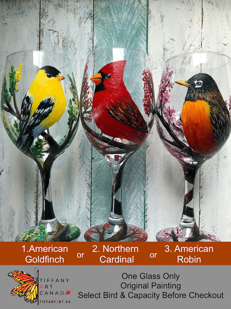 May include: Three hand-painted wine glasses, each featuring a different bird: an American Goldfinch, a Northern Cardinal, and an American Robin. The birds are depicted perched on branches with colorful foliage. The text at the bottom says "Select Bird & Capacity Before Checkout."