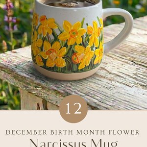May include: A ceramic mug featuring a hand-painted design of yellow narcissus flowers with green stems against a speckled white background. The mug is filled with a dark liquid and includes the text 12 DECEMBER BIRTH MONTH FLOWER Narcissus Mug.