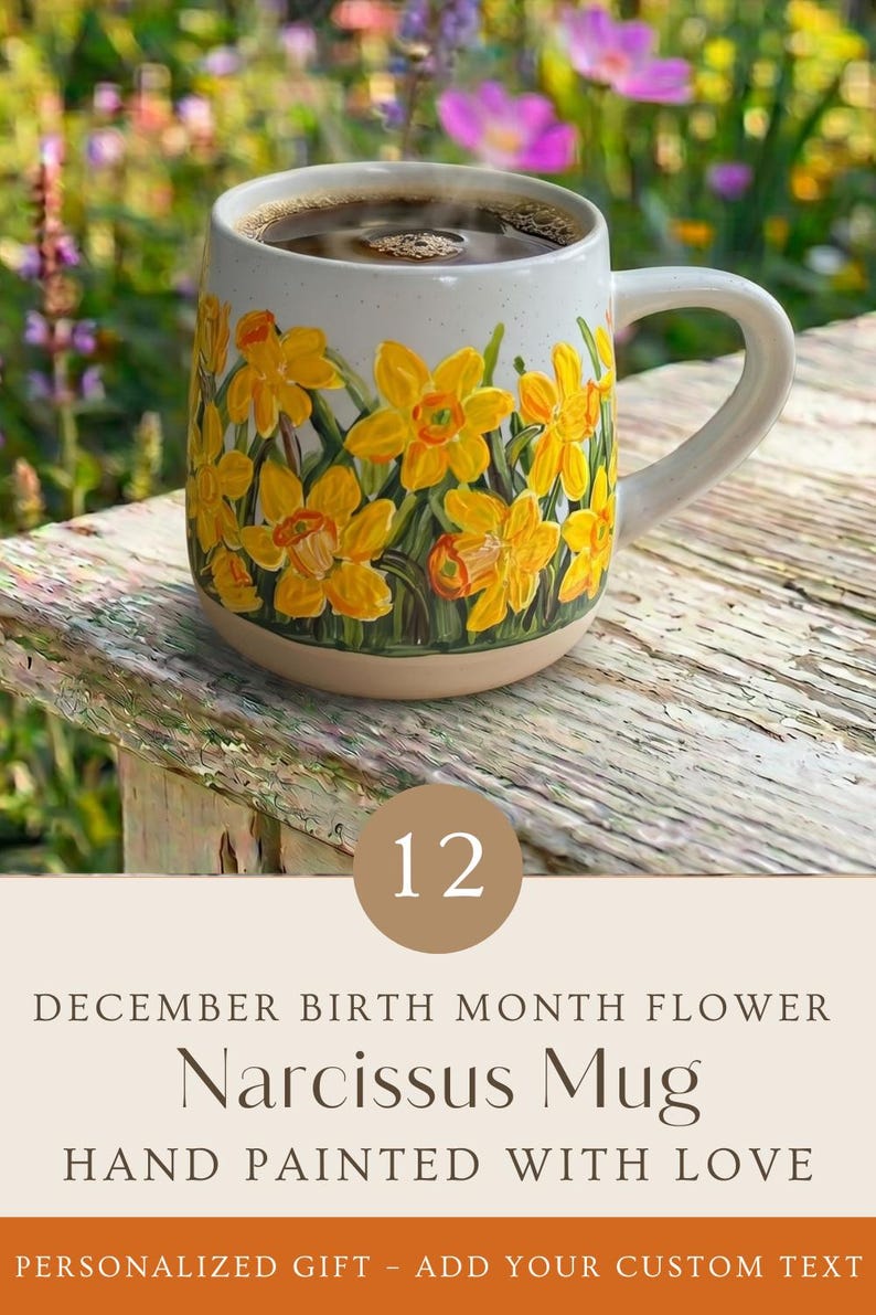 May include: A ceramic mug featuring a hand-painted design of yellow narcissus flowers with green stems against a speckled white background. The mug is filled with a dark liquid and includes the text 12 DECEMBER BIRTH MONTH FLOWER Narcissus Mug.