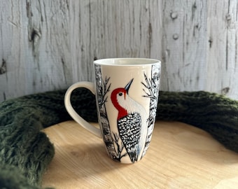 May include: White ceramic mug with a black and white illustration of a red-headed woodpecker on a birch tree background.