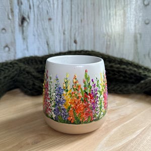 May include: A white ceramic tumbler featuring a hand-painted design of colorful lupine flowers in shades of orange, yellow, purple, pink, and green. The vessel has a rounded, stemless shape with a smooth finish and a tapered base.
