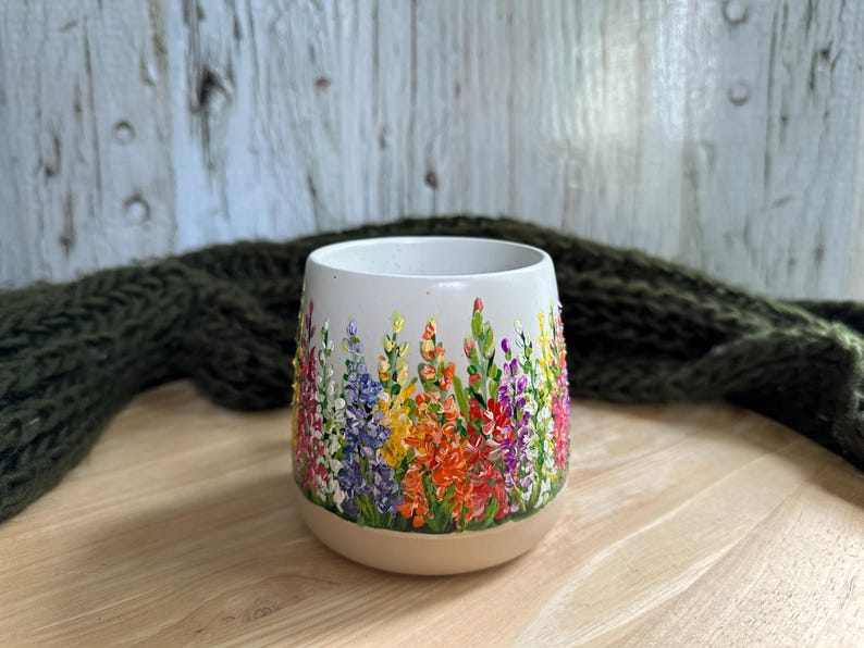 May include: A white ceramic tumbler featuring a hand-painted design of colorful lupine flowers in shades of orange, yellow, purple, pink, and green. The vessel has a rounded, stemless shape with a smooth finish and a tapered base.