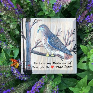 May include: A wooden memorial plaque featuring a hand-painted hawk perched on a branch. The plaque includes the text "In Loving Memory of Joe Smith 1941-2021" with a heart symbol. The background shows a natural wood grain.