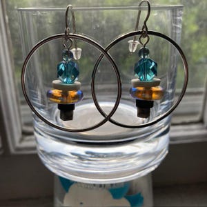 May include: A pair of hoop earrings with a silver finish. Each earring features a cluster of beads in shades of blue, white, gold, and black. The earrings are displayed in a clear glass.