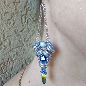 May include: A silver and blue beaded earring with a dangling chain. The earring features a silver ball stud surrounded by blue beads. The chain leads to a cluster of silver and blue elements, culminating in a teardrop-shaped bead with iridescent hues.