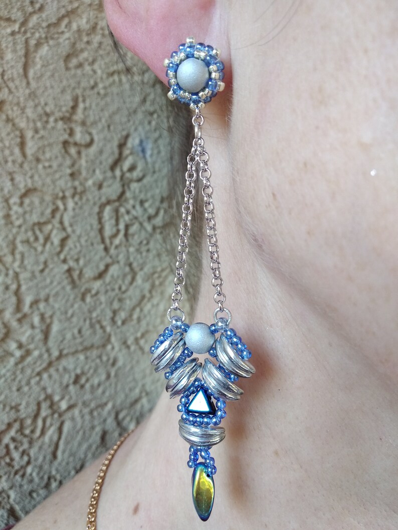 May include: A silver and blue beaded earring with a dangling chain. The earring features a silver ball stud surrounded by blue beads. The chain leads to a cluster of silver and blue elements, culminating in a teardrop-shaped bead with iridescent hues.
