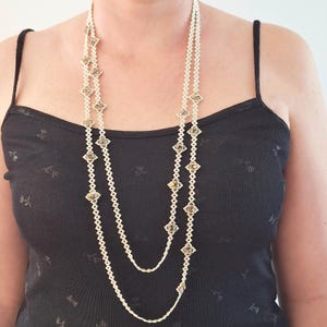 Puede incluir: Un collar largo de varias capas con cuentas blancas y detalles dorados en forma de diamante. El collar se lleva sobre una camiseta sin mangas negra. Las cuentas son pequeñas y espaciadas uniformemente, creando un aspecto delicado y elegante.