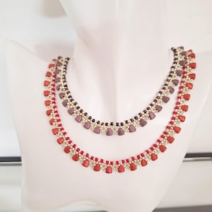 Op de afbeelding: Twee kettingen met kralen op een witte mannequin. De onderste ketting heeft rode hartvormige kralen, terwijl de bovenste ketting paarse hartvormige kralen heeft. Beide kettingen hebben zilverkleurige accenten en kleine kralen.