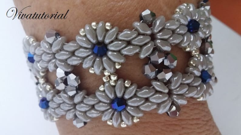 May include: A grey beaded bracelet with silver and blue accents. The bracelet is made with small, oval-shaped beads and features a repeating pattern of flower-like shapes.