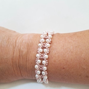 May include: A beaded bracelet featuring rows of white pearls and rose gold beads. The bracelet is designed with a repeating pattern of pearl clusters, creating a delicate and elegant look. The bracelet is shown on a person's wrist.