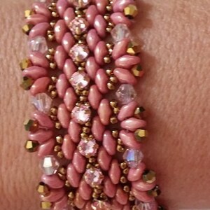 Puede incluir: Un brazalete de cuentas rosa y dorado con un patr&oacute;n tejido. El brazalete est&aacute; hecho con peque&ntilde;as cuentas rosas y cuentas doradas, y presenta detalles de cristal transparente.