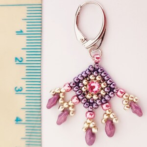 K&ouml;nnte beinhalten: Ein Paar H&auml;ngeohrringe mit einem Design aus lila, rosa und goldenen Perlen. Die Ohrringe haben einen kleinen rosa Kristall in der Mitte des Designs.