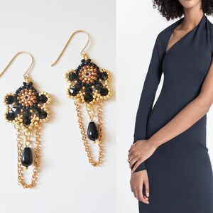 K&ouml;nnte beinhalten: Ein Paar goldene Ohrringe mit schwarzen und goldenen Perlen und einem schwarzen Tropfen-Anh&auml;nger. Die Ohrringe sind baumelnd und haben ein florales Design.