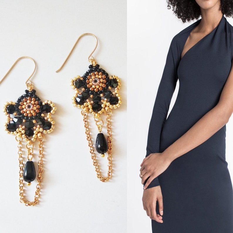 K&ouml;nnte beinhalten: Ein Paar goldene Ohrringe mit schwarzen und goldenen Perlen und einem schwarzen Tropfen-Anh&auml;nger. Die Ohrringe sind baumelnd und haben ein florales Design.